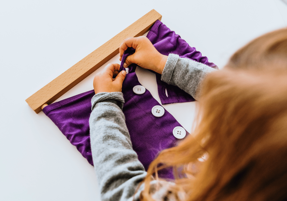 Дете упражнява фината си моторика чрез закопчаване на бели копчета върху лилав плат.