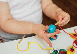 Дете тренира концентрацията си чрез низане на големи сини мъниста върху връв.